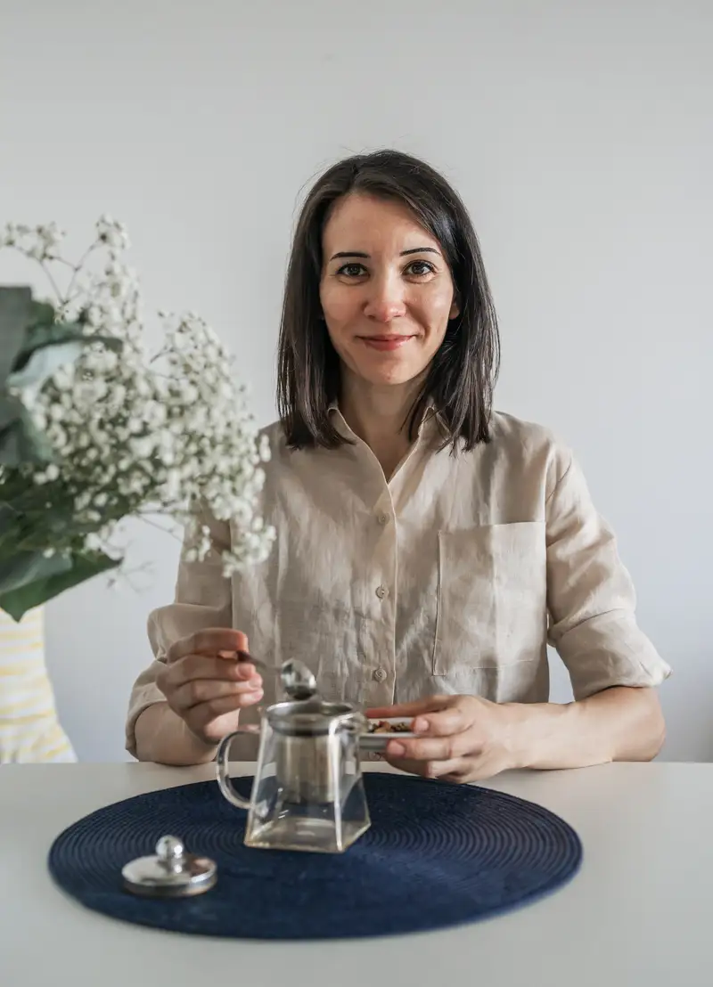 Porträt einer lächelnden Frau am Tisch beim Teezubereiten mit einer Glaskanne und einem Dosierlöffel