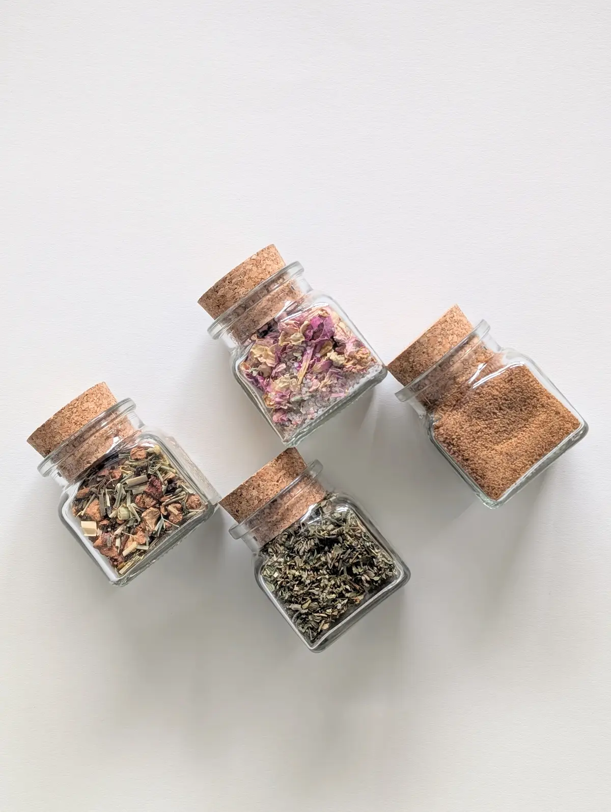 Top-down view of small square glass jars with cork lids and assorted loose teas on a white background